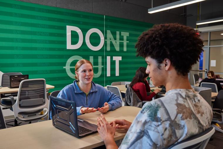 Two students talking across a table with a laptop nearby