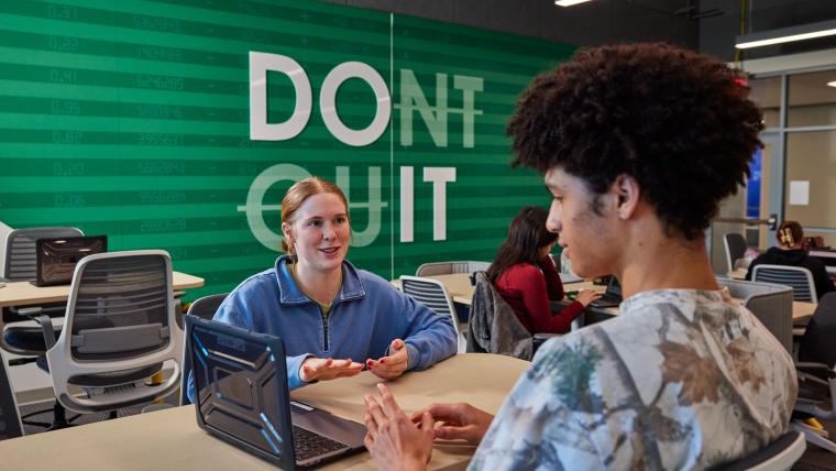Two students talking across a table with a laptop nearby
