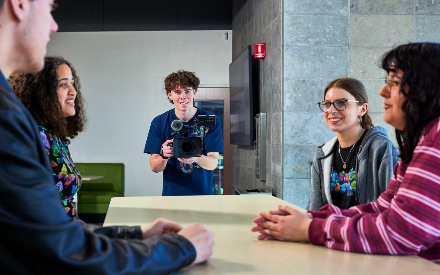 A male student taking video of a group of student sitting at a table.