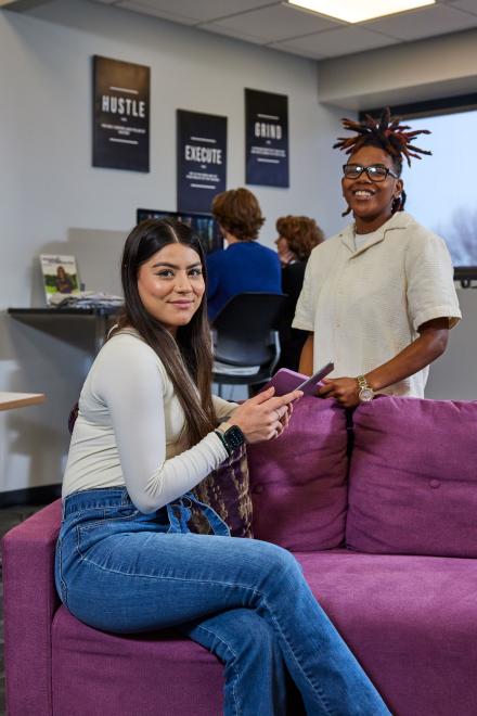 Student sitting on a couch visiting with a student standing.